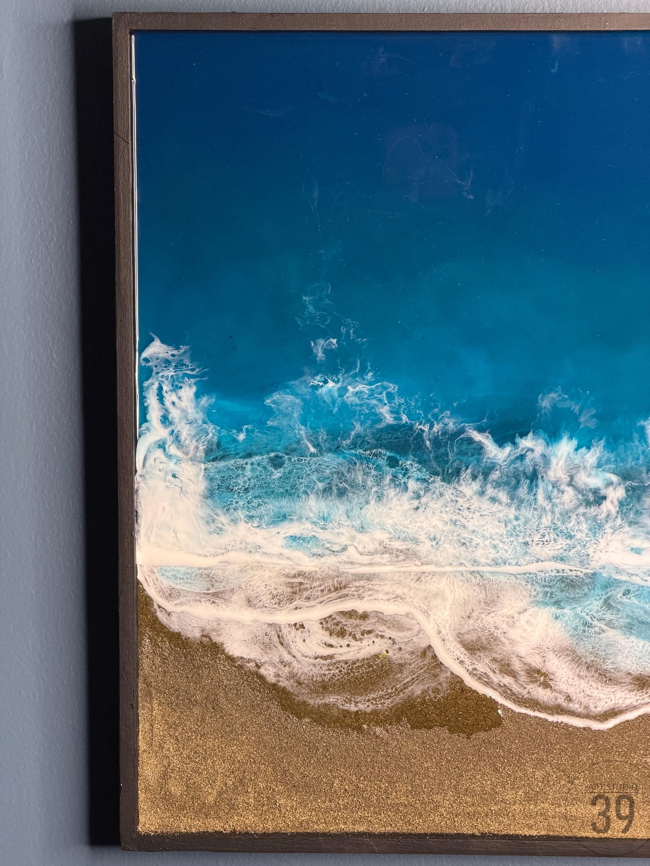 Ocean-themed artwork featuring a vibrant depiction of a beach scene. The artwork showcases a deep blue ocean with foamy white waves crashing onto a golden sandy shore. The piece is framed in a dark brown wooden frame.