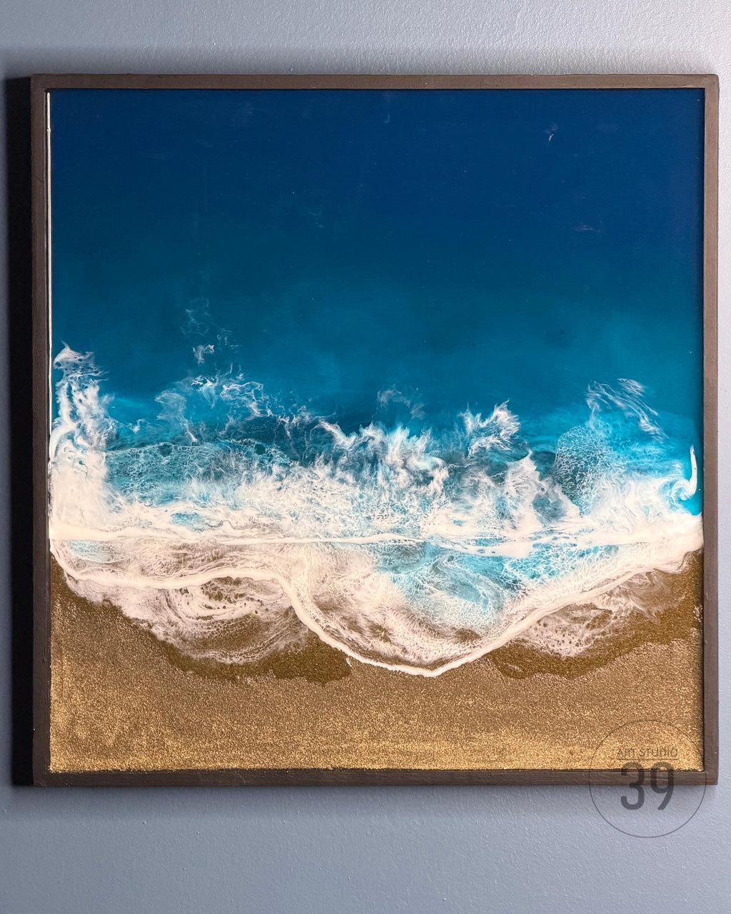 Square framed artwork depicting an aerial view of a beach scene. The top portion is a deep blue, transitioning to turquoise and white waves. The bottom is a golden sand effect. The artwork is framed in a dark brown wood.