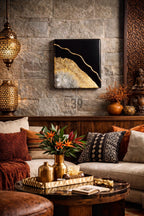 A square black and gold abstract art piece hangs on a stone wall. The artwork features a black background with gold and silver accents. The room is decorated with a sofa, pillows, and a coffee table, creating a warm and inviting atmosphere.