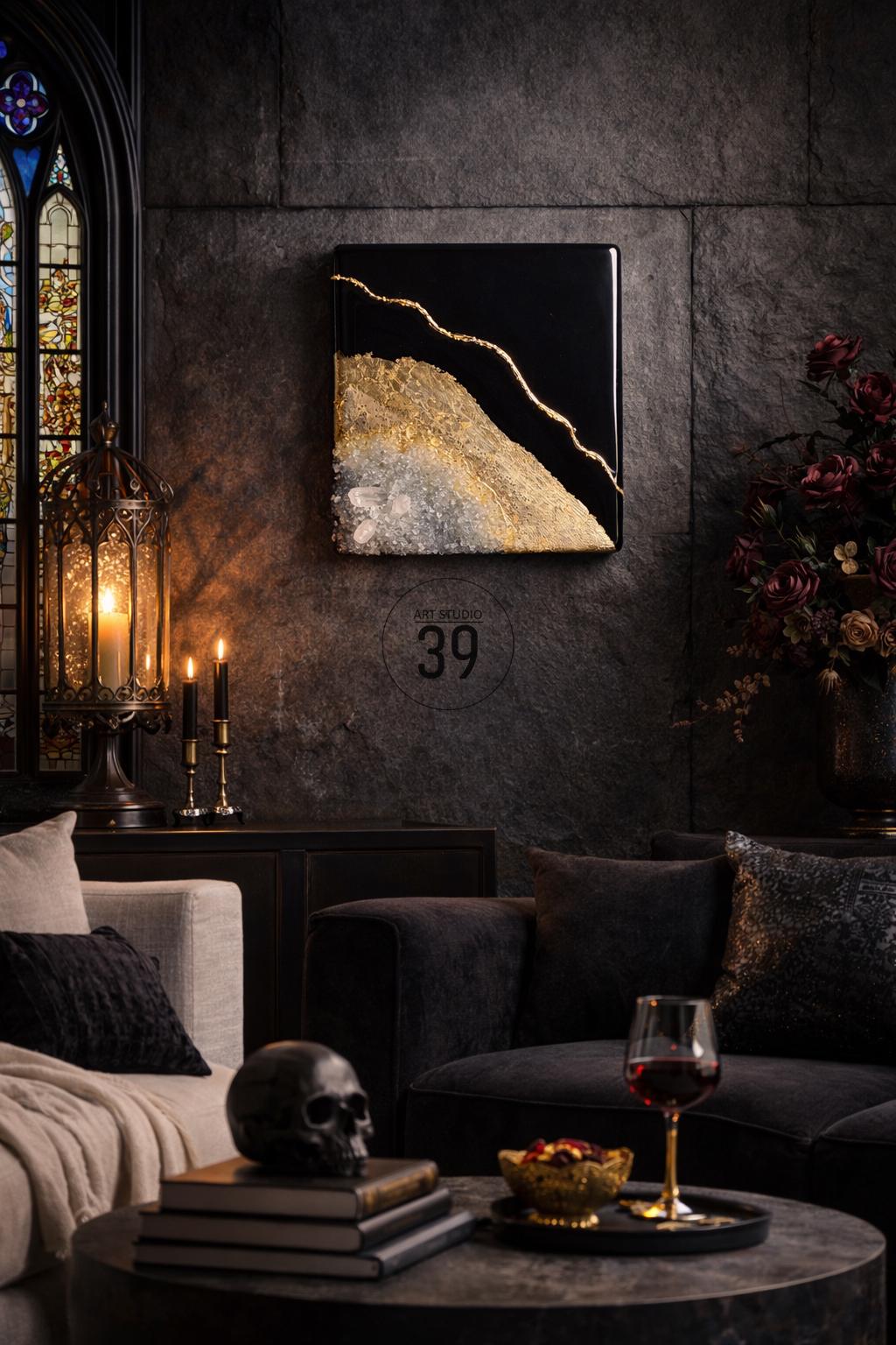 A square black and gold abstract art piece hangs on a dark gray stone wall. The artwork features a gold line separating black and gold textured sections. The room is decorated with a black skull, candles, and a dark gray sofa.