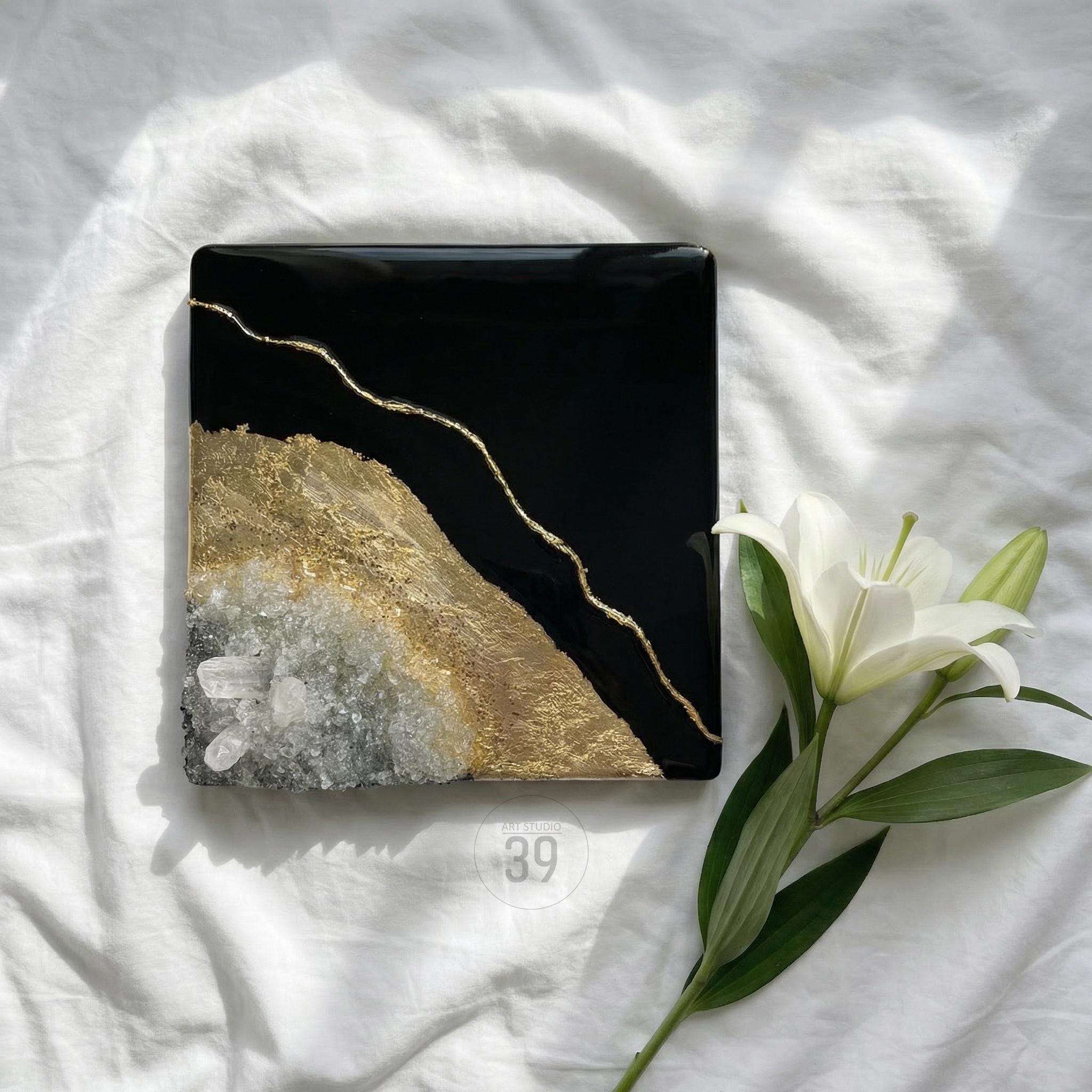 A square, black resin art piece with gold accents and a textured, gold and white section. A gold line runs diagonally across the black surface. The number "39" is visible. A white lily flower is in the image.