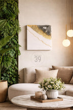 A modern living room with a white sofa, decorative pillows, and a round coffee table. A square abstract art piece with gold and gray accents hangs on the wall. The image also features a vertical garden and pendant lights.