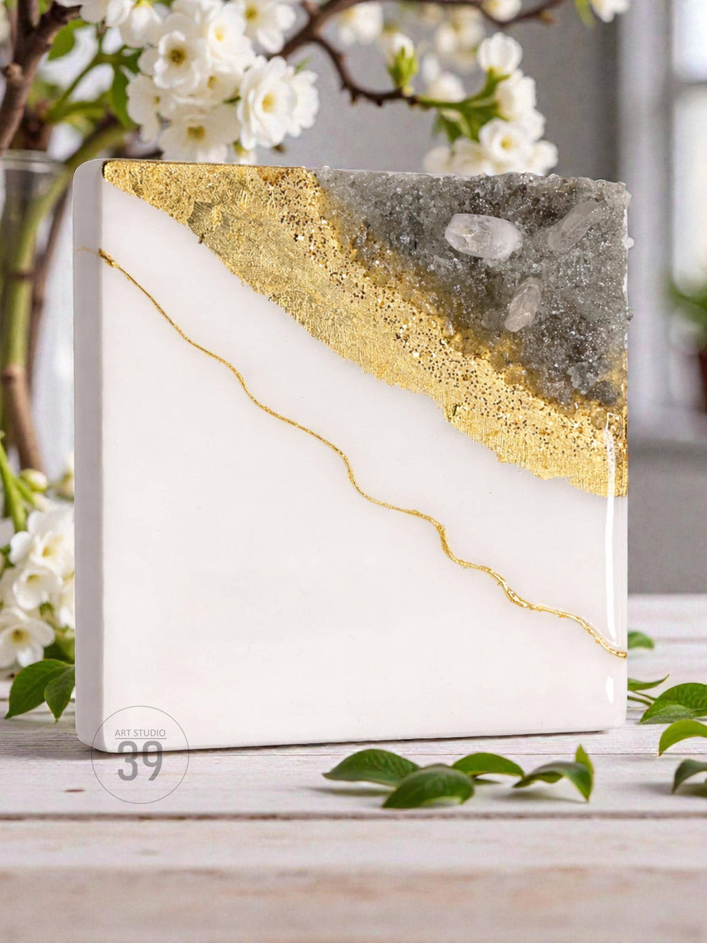A square, white resin art piece with a diagonal gold leaf and gray crystal design. A thin gold line curves across the white surface. The text "ART STUDIO 39" is visible on the bottom left corner.