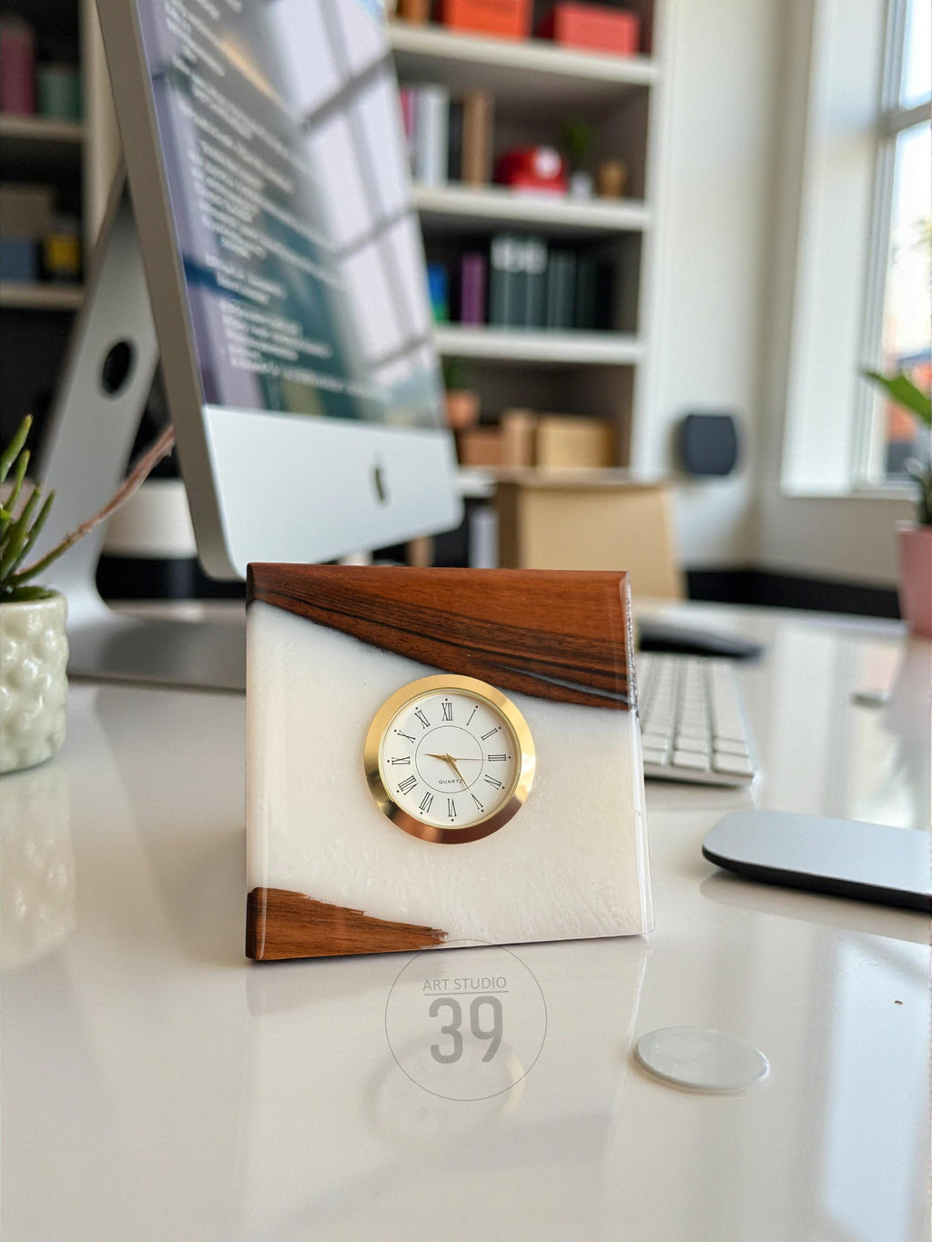 A square clock with a white face and gold-colored trim. The clock is set in a white resin block with wood accents. The clock face displays Roman numerals. The text "ART STUDIO 39" is visible.
