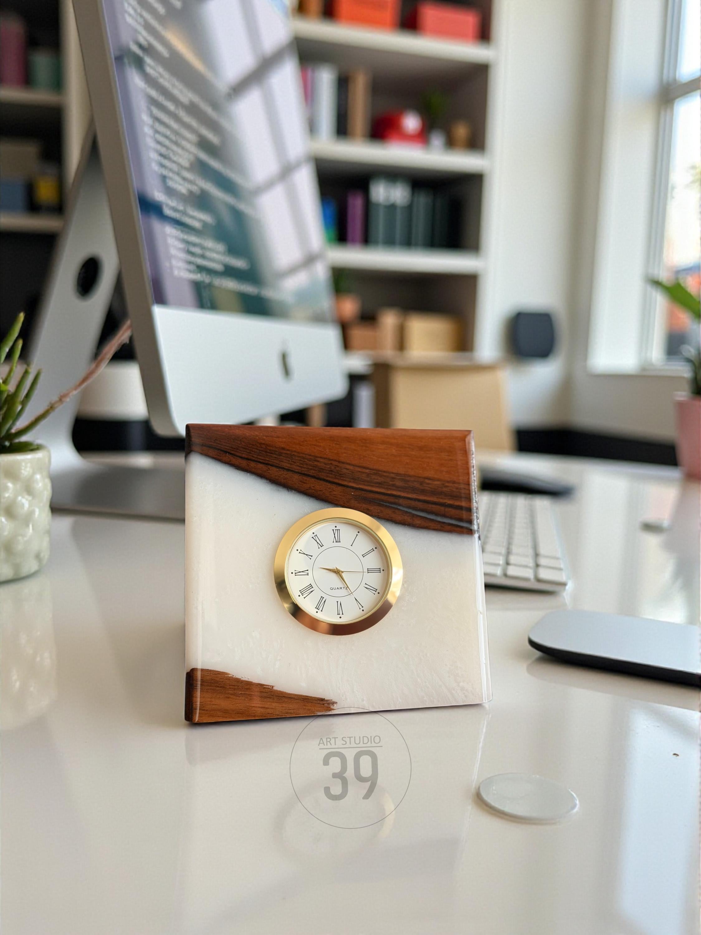 A square clock with a white face and gold-colored trim. The clock is set in a white resin block with wood accents. The clock face displays Roman numerals. The text "ART STUDIO 39" is visible.