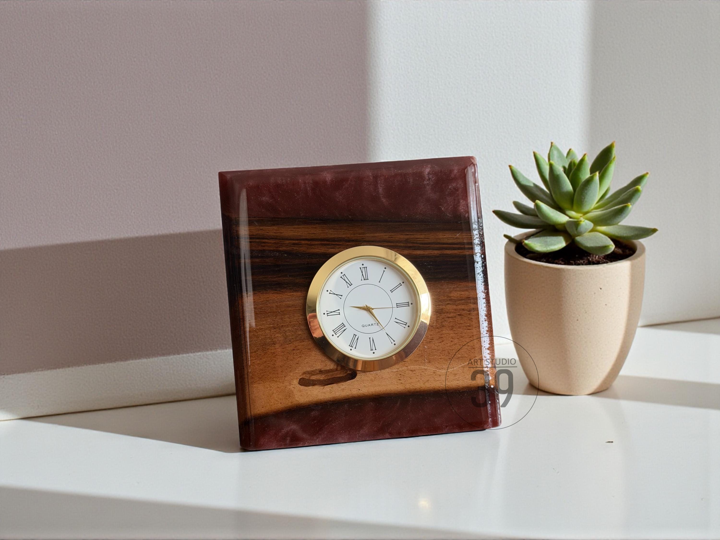 A square wooden clock with a gold-colored clock face and hands. The clock face has black Roman numerals. The wood has a dark brown and reddish-brown resin finish. A small succulent plant in a beige pot is next to the clock.