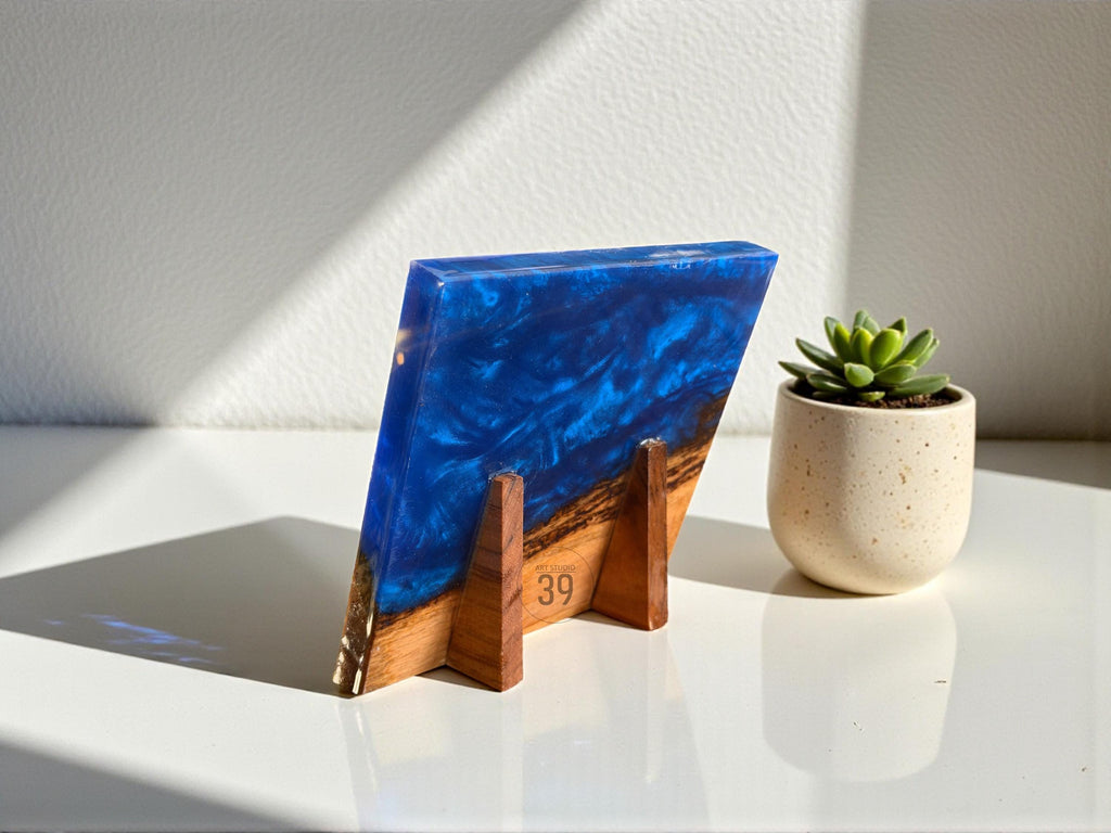 A rectangular resin art piece with a deep blue and white swirling design, supported by a wooden stand. The piece is angled, revealing a glimpse of a wooden base with the number "39" visible. A small potted succulent sits nearby.