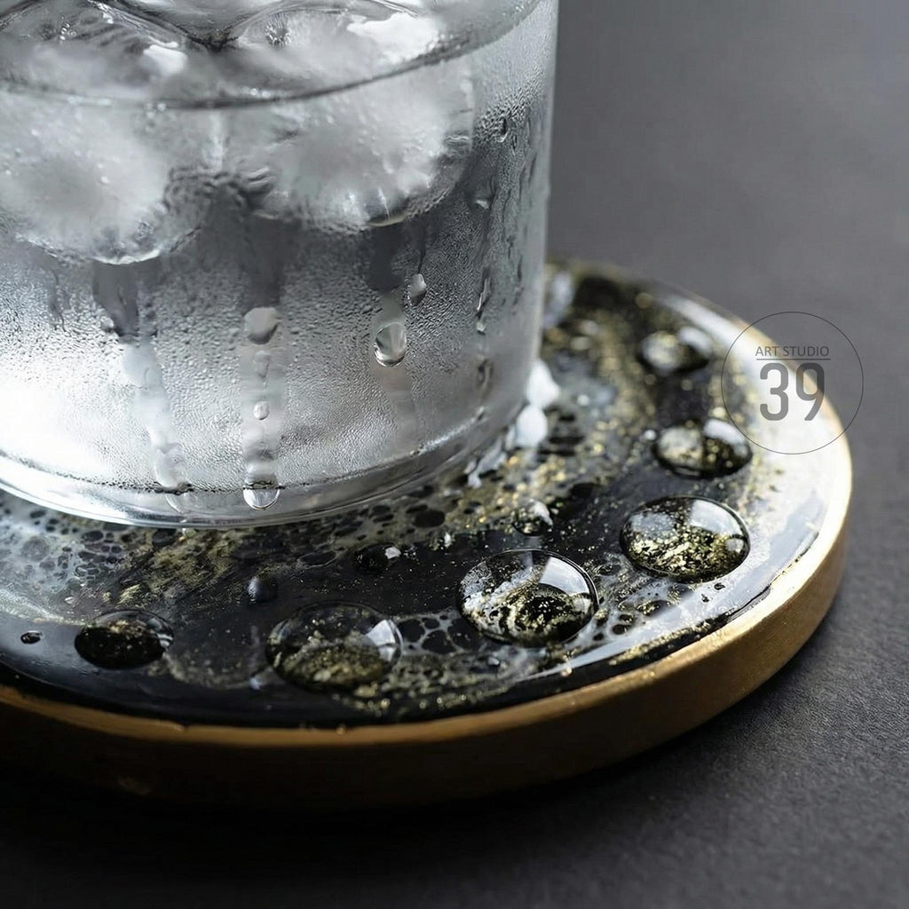 A clear glass filled with ice and water sits on a black and gold coaster. The coaster has a textured surface with gold accents and a gold rim. Water droplets are visible on the coaster and glass. The text "ART STUDIO 39" is visible.