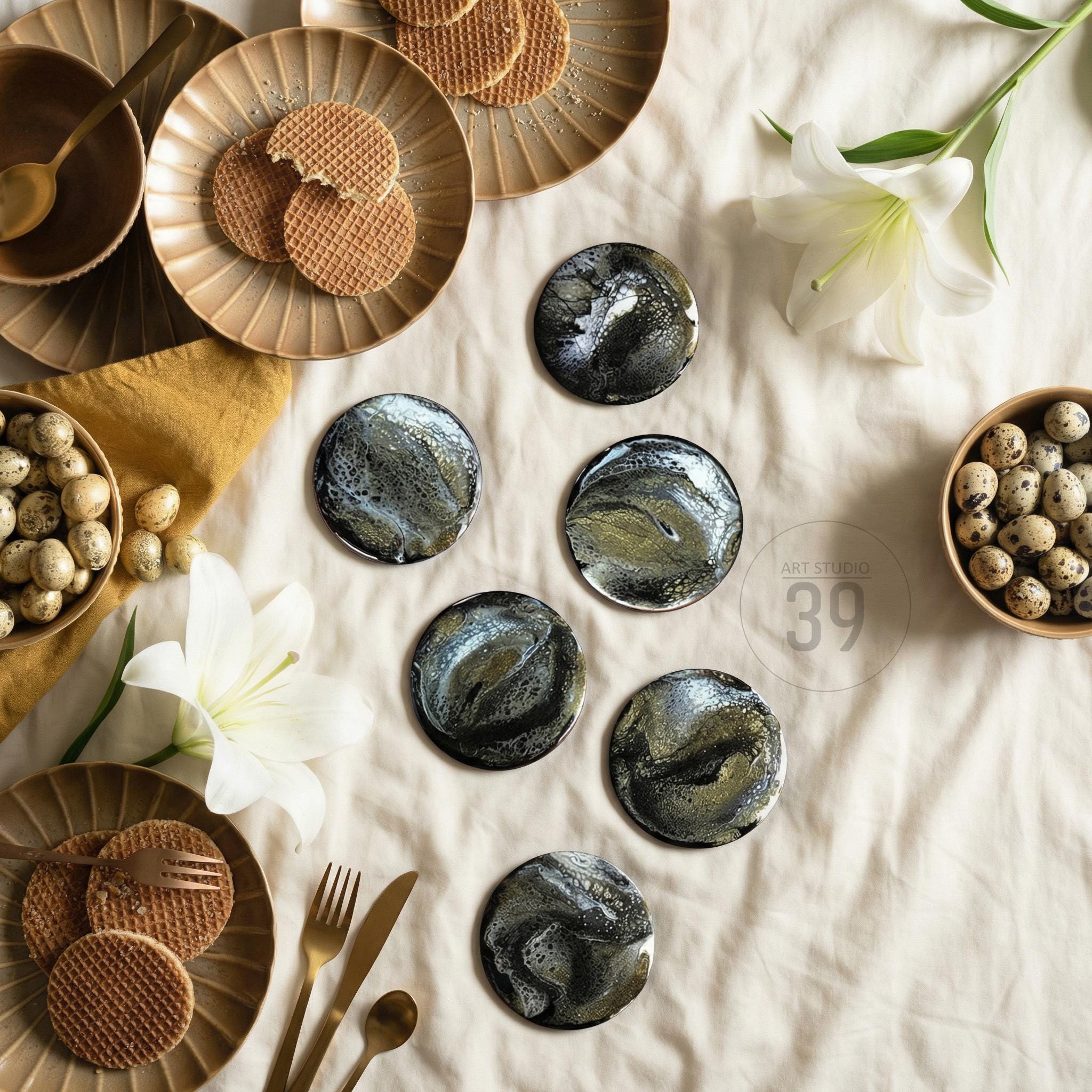 Six round coasters with a black, white, and gold abstract design are arranged on a white surface. The coasters are surrounded by plates of cookies, bowls of eggs, and gold utensils. The image also includes the text "ART STUDIO 39".
