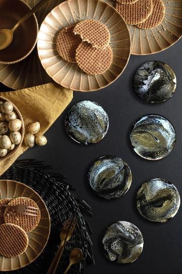 An overhead shot features a collection of decorative coasters with a marbled design in black, white, and gold. Waffle cookies and plates are also present, along with a bowl of eggs and serving utensils, all arranged on a black surface.