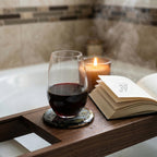 A stemless wine glass filled with red wine sits on a coaster atop a wooden bath tray. A lit candle and an open book with the text "ART STUDIO 39" are also on the tray, creating a relaxing spa-like atmosphere.