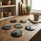 Six round coasters with a marbled design in black, gold, and white are arranged on a wooden surface. The coasters have a glossy finish and are placed near a mug and a small potted plant. The scene suggests a kitchen or dining setting.