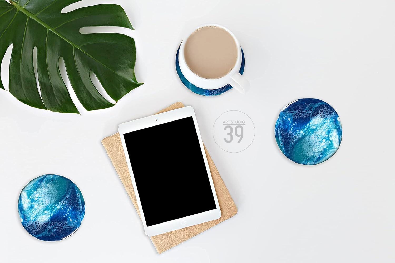 An overhead shot of a white surface with a coffee cup, a tablet, and three blue and white coasters. A large green leaf is in the upper left corner. The coasters have a swirling blue and white design. The tablet has a black screen. The text "ART STUDIO 39" is in a circle.