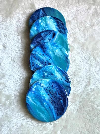 A stack of six round coasters with a blue and turquoise marbled design. The coasters have a glossy finish and are arranged vertically, showcasing the fluid patterns of the paint. The coasters are on a white, textured surface.