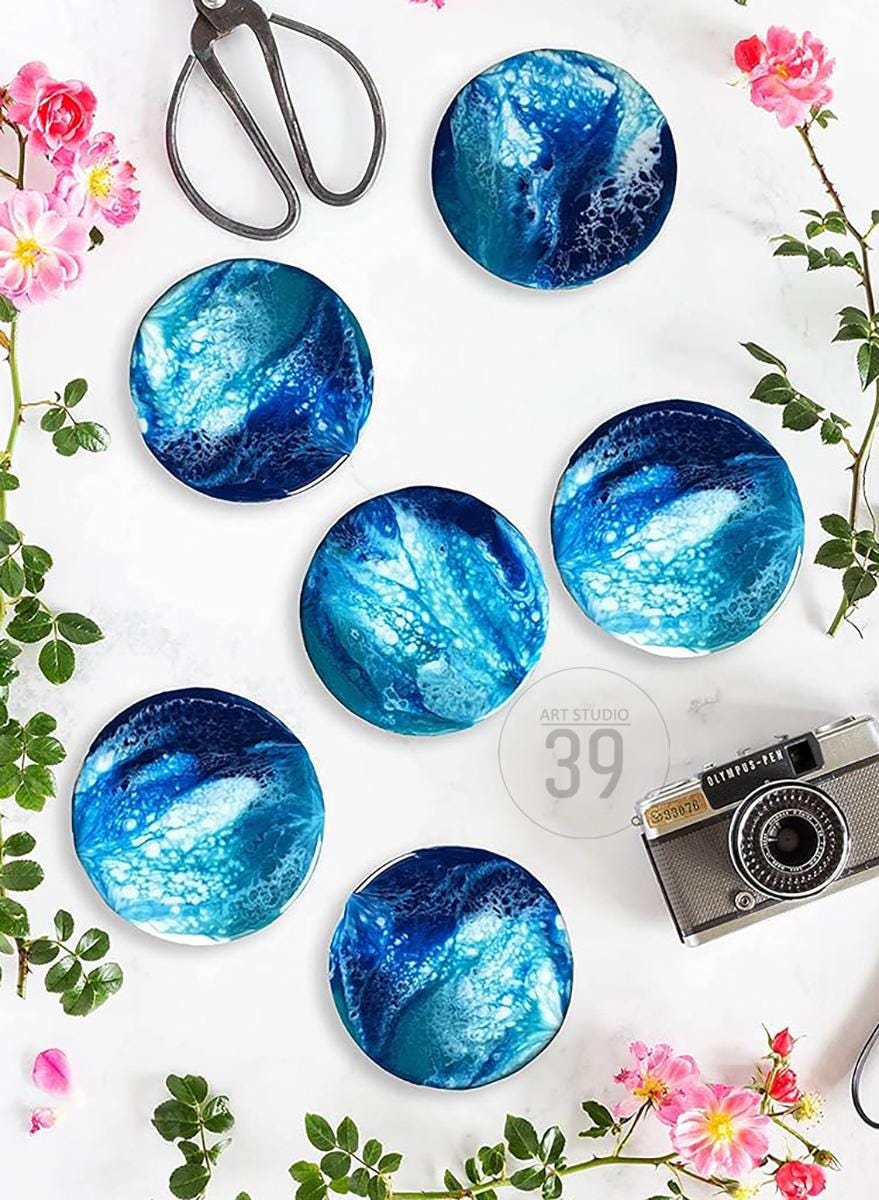 Six round coasters with a blue and white abstract design, resembling ocean waves. The coasters are arranged on a white surface, with pink flowers, scissors, and a vintage camera. The text "ART STUDIO 39" is visible.