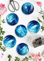 Six round coasters with a blue and white abstract design, resembling ocean waves. The coasters are arranged on a white surface, with pink flowers, scissors, and a vintage camera. The text "ART STUDIO 39" is visible.