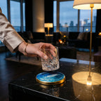 A clear, patterned glass with the number 39 on it is held above a blue and gold coaster. The coaster has a marbled design. The scene is set in a modern living room with a city view.