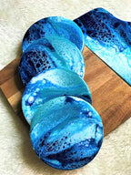 A set of four round coasters and a rectangular serving board. The coasters and board feature a blue and white resin design, resembling ocean waves. The coasters are arranged on a wooden surface.
