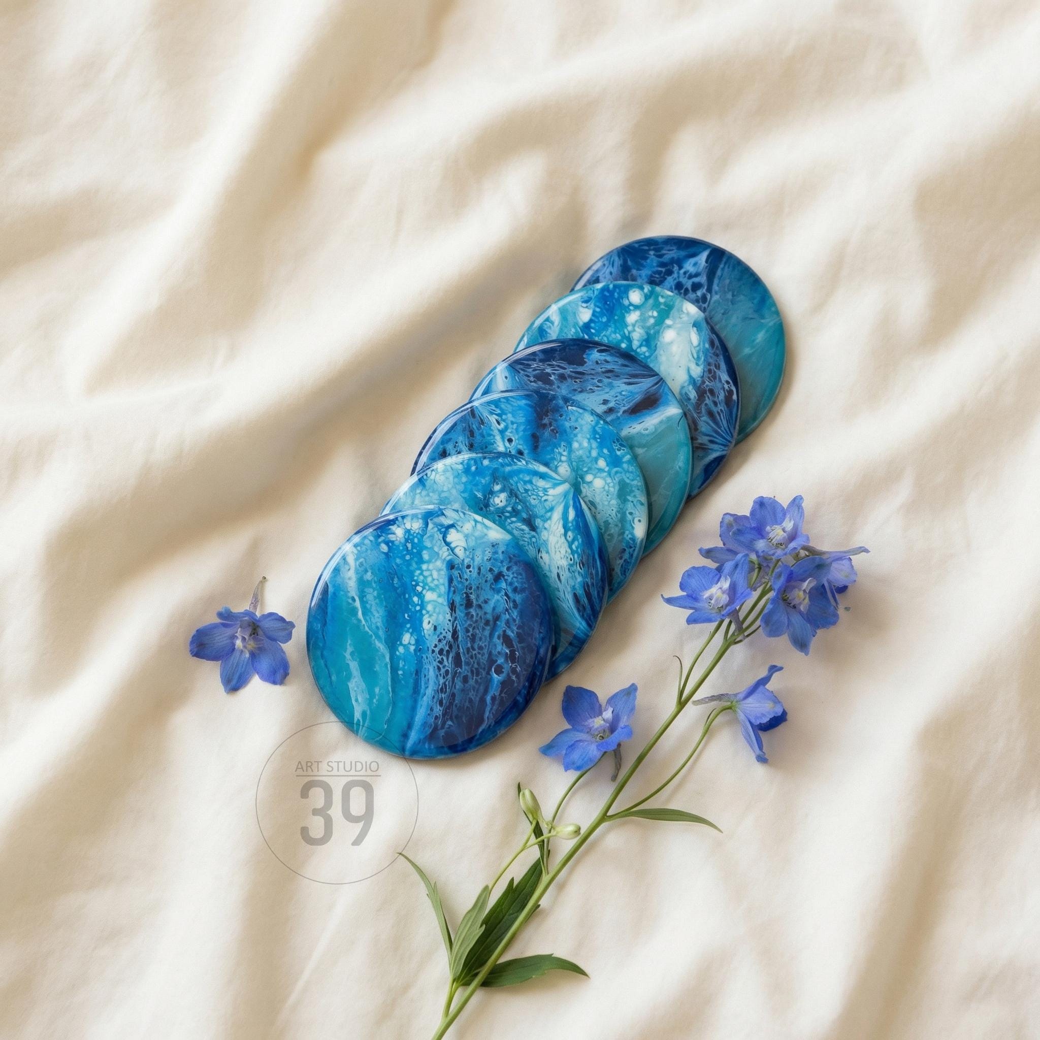 A set of six round coasters with a blue and white abstract design, arranged on a white fabric surface. The coasters have a marbled effect, with shades of blue. Blue flowers and the text "ART STUDIO 39" are also visible.