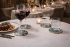 A table setting with two wine glasses filled with red wine, two glasses of water, and a plate of food. The glasses sit on blue and gold coasters. A lit candle and silverware are also present.