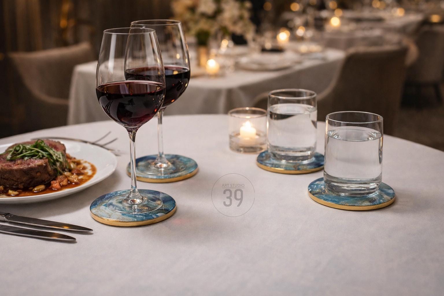 A table setting with two wine glasses filled with red wine, two glasses of water, and a plate of food. The glasses sit on blue and gold coasters. A lit candle and silverware are also present.