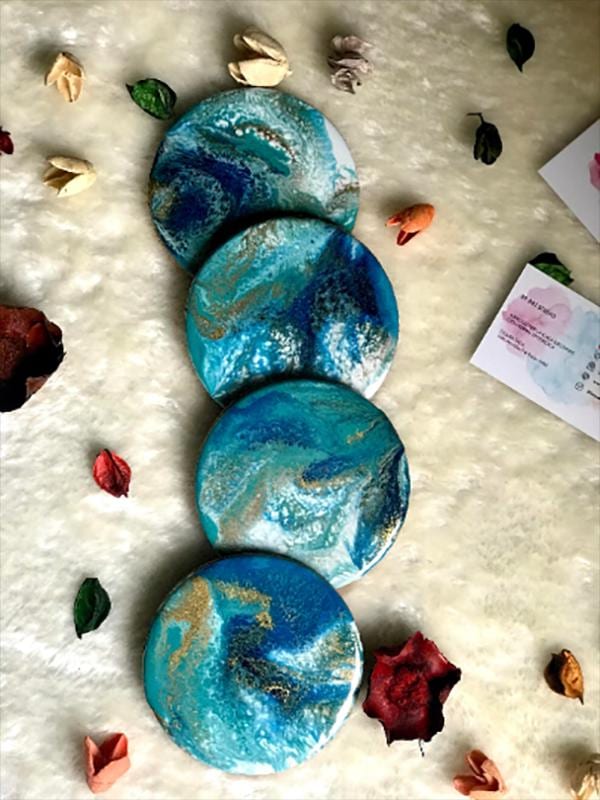 Four round coasters with a blue, teal, and gold swirl design. The coasters are arranged in a vertical stack on a white, textured surface, with dried flowers and leaves scattered around.