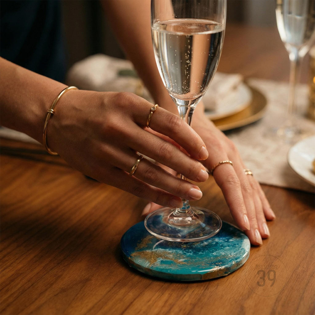 A person holding a champagne flute above a blue and gold resin coaster. The coaster is on a wooden table. The person is wearing gold rings and a gold bracelet.