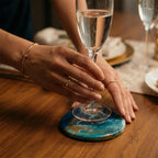 A person holding a champagne flute above a blue and gold resin coaster. The coaster is on a wooden table. The person is wearing gold rings and a gold bracelet.