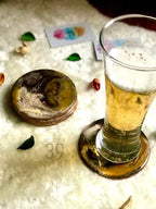 A stack of round coasters with a marbled design in shades of brown, gold, and white. A glass of beer sits on one coaster. The coasters are next to a card with colorful watercolor splotches. The text "ART STUDIO 39" is visible.