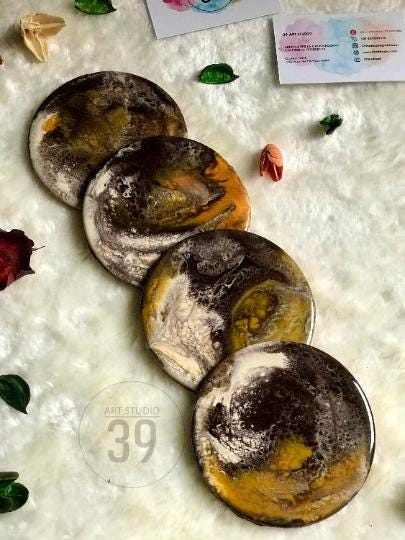 Four round coasters with a marbled design in shades of brown, yellow, and white. The coasters are arranged diagonally on a white, fluffy surface. The text "ART STUDIO 39" is visible.