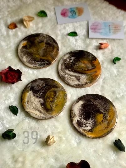 Four round coasters with abstract designs in shades of brown, gold, and white. The coasters are arranged on a textured white surface, with small decorative elements scattered around. The text "ART STUDIO 39" is visible.