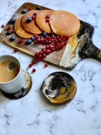A wooden serving board with pancakes, blueberries, and red currants. A white mug of coffee sits next to a matching coaster. A second coaster is also visible. The board has a decorative handle.