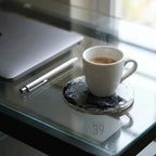A white ceramic coffee cup with a dark liquid sits on a black and gray coaster. A silver pen and a laptop are on a glass surface. The coaster has a marbled design. The text "ART STUDIO 39" is visible.