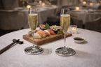 Two champagne flutes filled with sparkling wine sit on a table, next to a wooden board of sushi and a small bowl of soy sauce. The flutes rest on decorative coasters. Chopsticks are placed on the table. The scene is set in a restaurant.