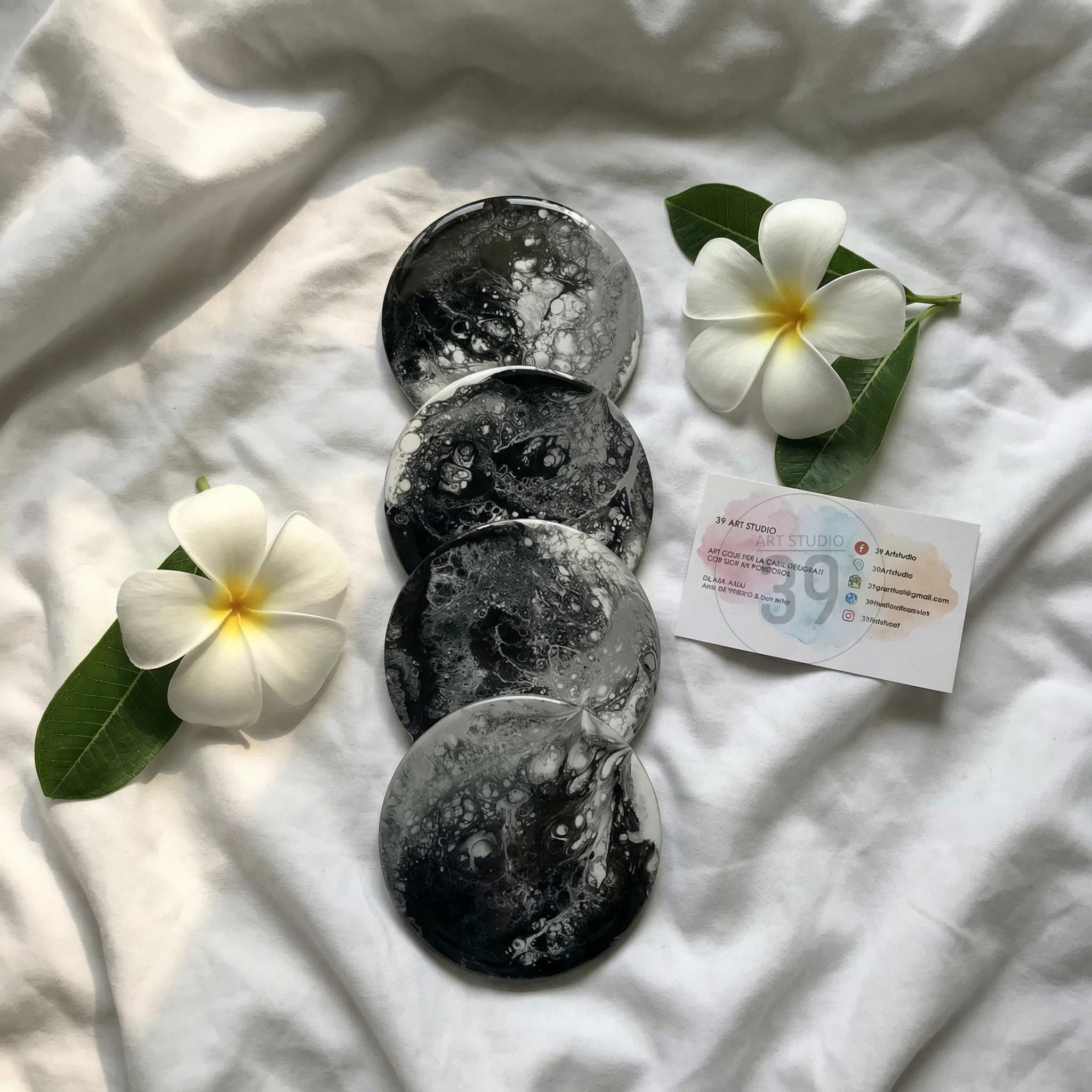 Four round coasters with a black and white abstract design. The coasters are arranged in a vertical stack on a white surface. Two white plumeria flowers and a business card are also present.