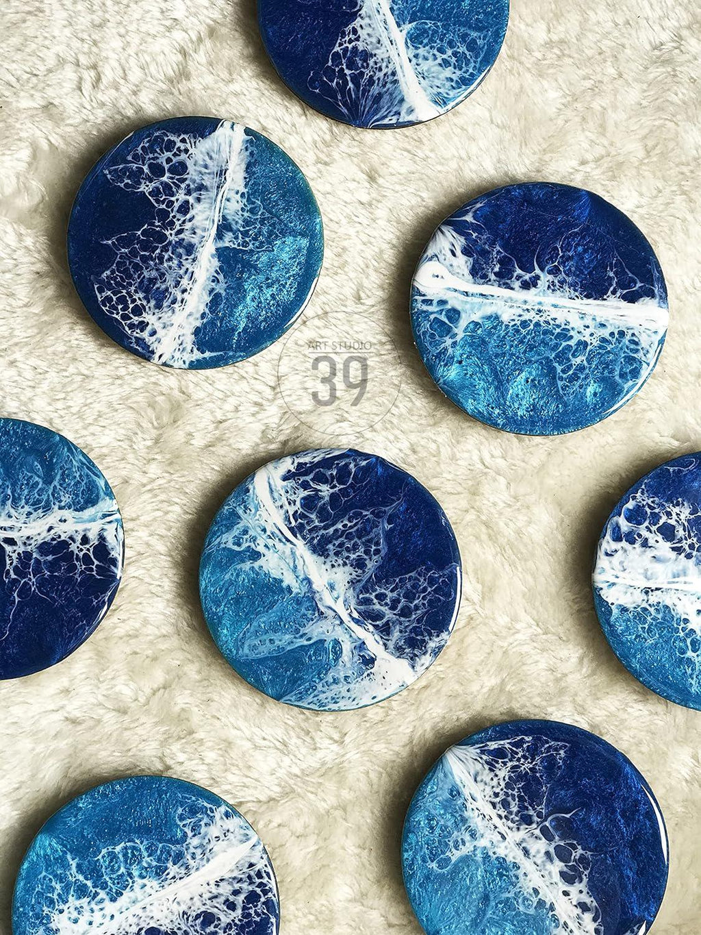 A collection of round coasters with an ocean-inspired design. Each coaster features a blend of deep blue, light blue, and white resin, creating a wave-like pattern. The coasters are arranged on a soft, white surface.