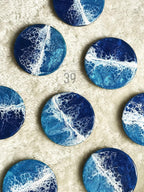 A collection of round coasters with an ocean-inspired design. Each coaster features a blend of deep blue, light blue, and white resin, creating a wave-like pattern. The coasters are arranged on a soft, white surface.