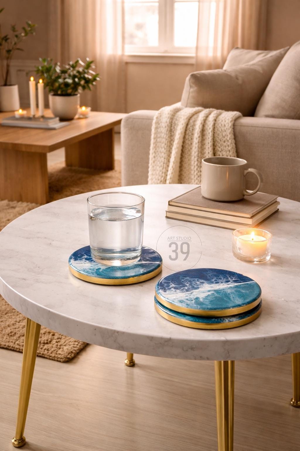 Two round coasters with a blue and white wave design and gold trim are stacked on a white marble table. A glass of water sits on one coaster. The text "ART STUDIO 39" is on the coaster. A lit candle and a mug are also on the table.