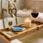 A wooden bathtub tray holds a lit scented candle, a wine glass with red wine, and a blue and white coaster. The tray is set over a bathtub with a gold faucet and white towels in the background. The coaster has a wave design.