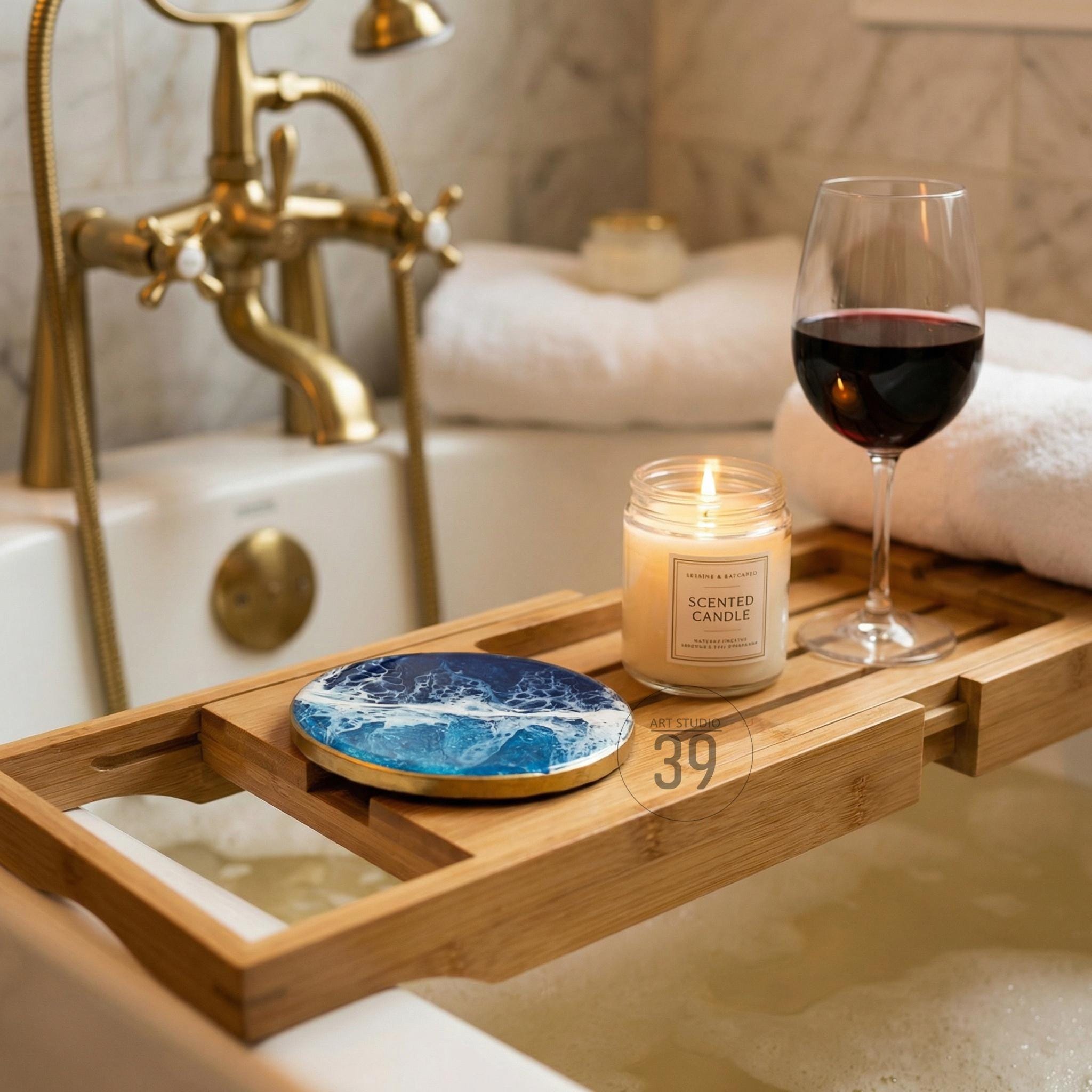 A wooden bathtub tray holds a lit scented candle, a wine glass with red wine, and a blue and white coaster. The tray is set over a bathtub with a gold faucet and white towels in the background. The coaster has a wave design.