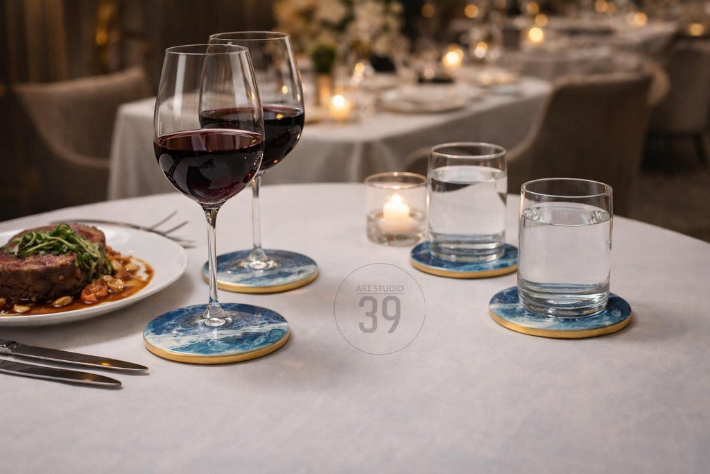 A table setting with two wine glasses filled with red wine, two glasses of water, and a plate of food. The coasters have a blue and white wave design with a gold edge. The scene is set in a restaurant.