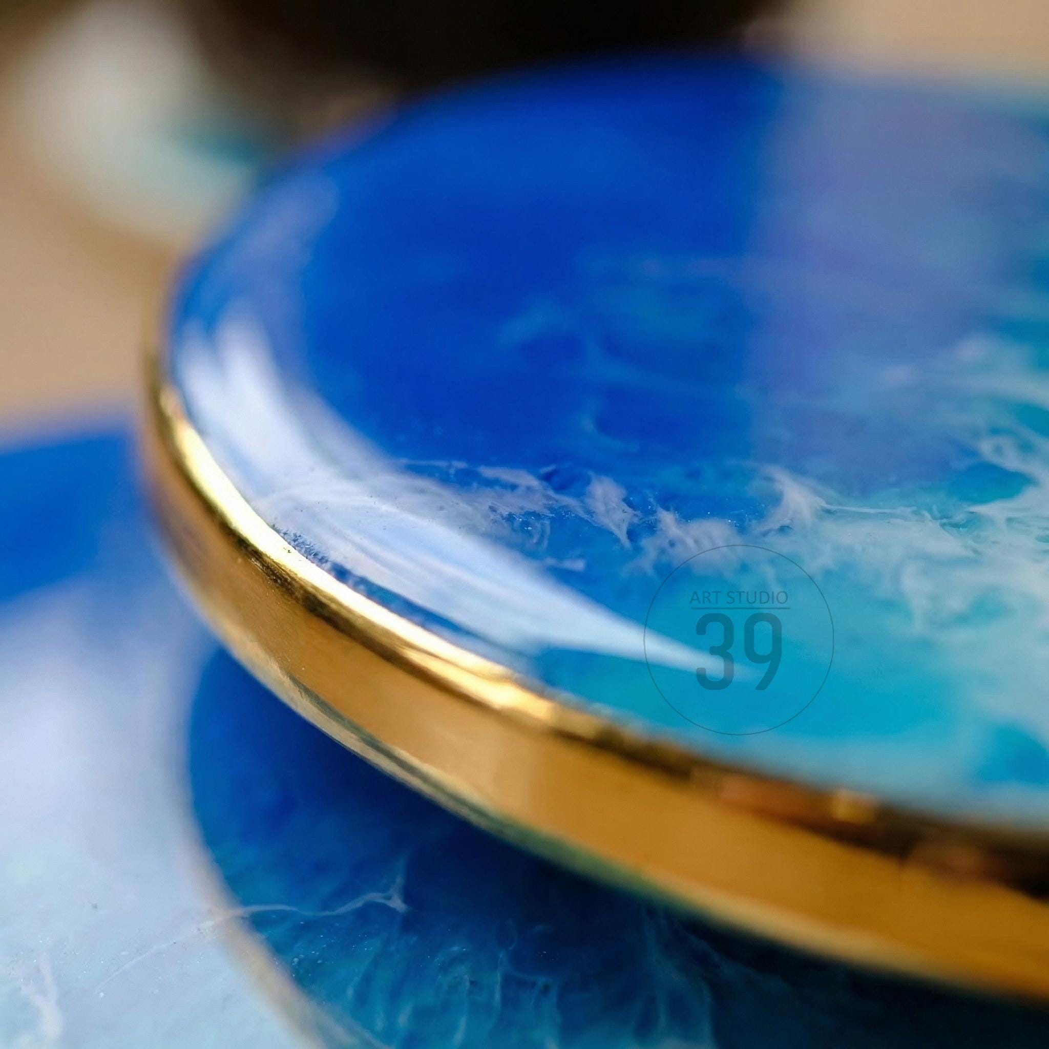 Close-up of a round, blue resin coaster with a gold rim. The coaster has a marbled white and blue design, resembling ocean waves. The text "ART STUDIO 39" is visible.