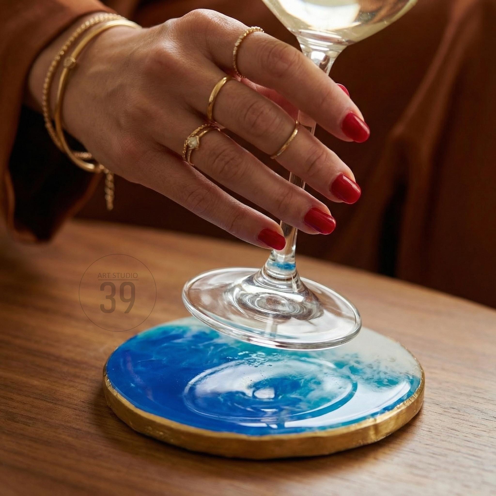 A round coaster with a blue and white ocean wave design, edged with gold. The coaster is under a wine glass. The image also shows a hand with gold rings and bracelets.