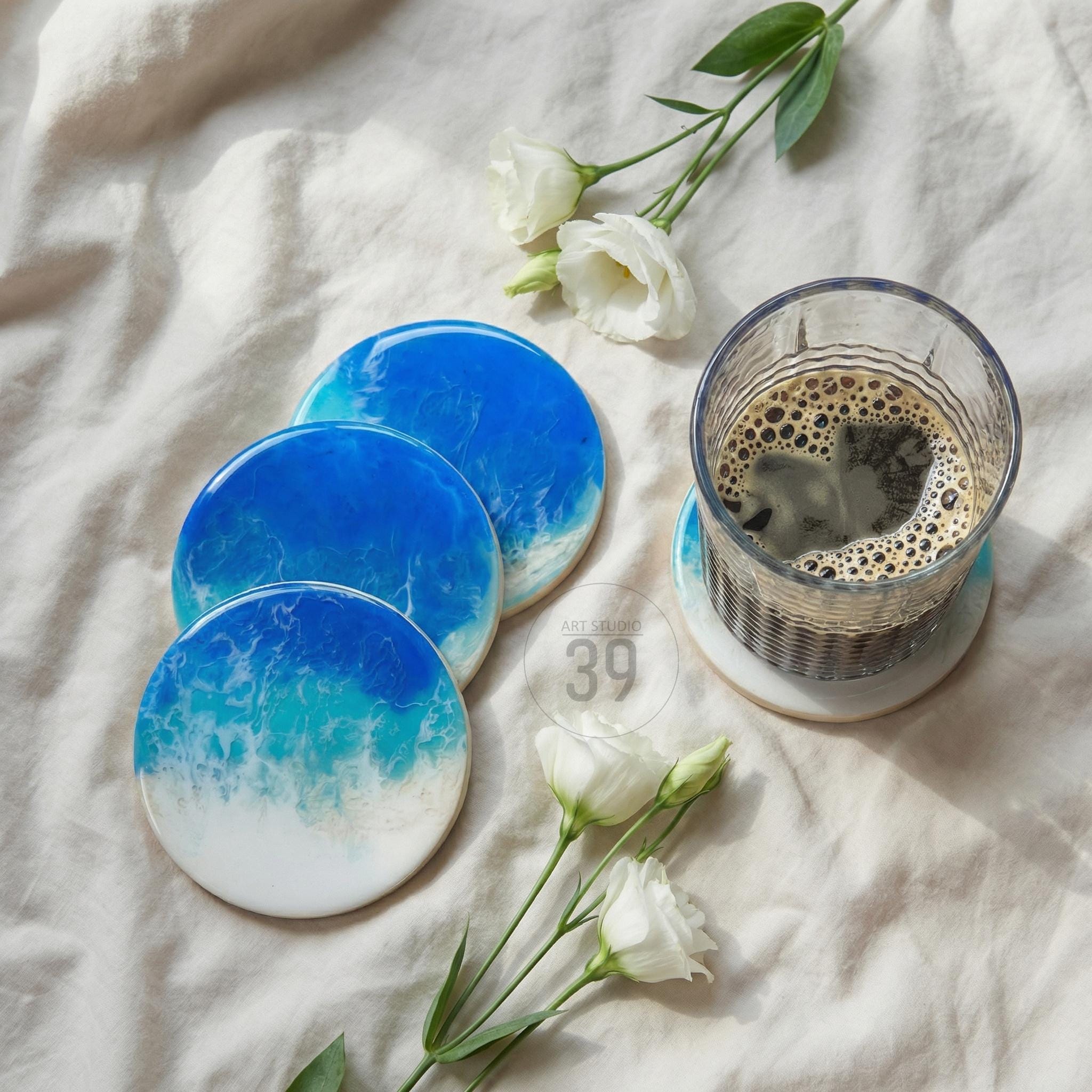 Set of four round coasters with a blue and white ocean wave design. A glass of coffee sits on one coaster. White flowers and the text "ART STUDIO 39" are also in the image.