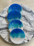 Four round coasters with a blue and white ocean wave design. The coasters are arranged on a marble-patterned tray. The coasters have a glossy finish and a beach-like appearance. The text "ART STUDIO 39" is visible.