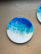 Round coaster with an ocean wave design. The coaster features shades of blue, turquoise, and white, resembling a seascape. The coaster is a decorative item with a glossy finish. The text "ART STUDIO 39" is printed on the coaster.