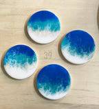 Four round coasters with a resin ocean wave design. The coasters feature a gradient of blue and turquoise hues, transitioning to white, resembling ocean foam. The text "ART STUDIO 39" is visible.