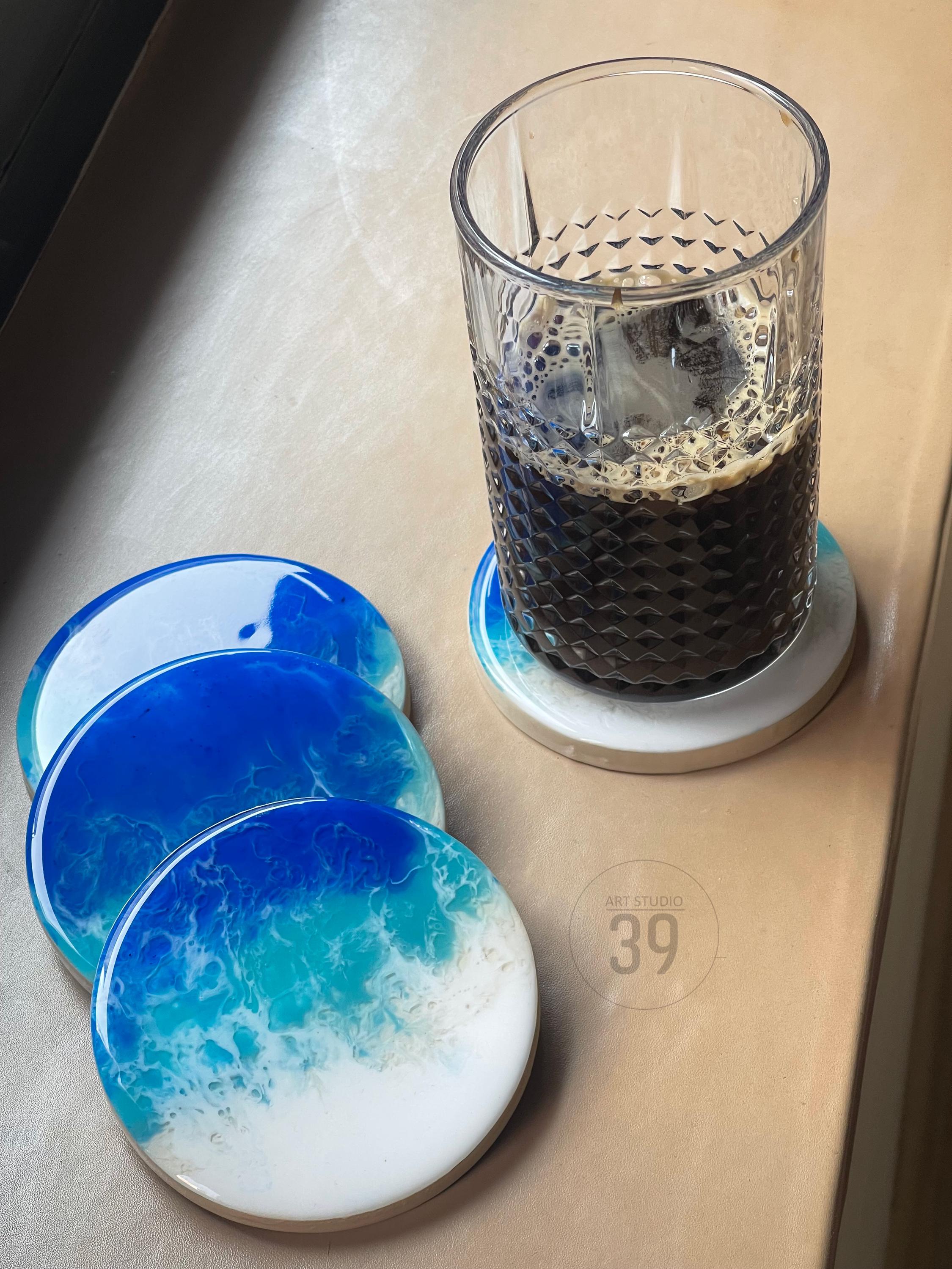 A clear glass of dark liquid sits on a round coaster with a blue and white ocean wave design. Several similar coasters are arranged nearby. The coasters have a glossy finish and are made of resin. The image includes the text "ART STUDIO 39".