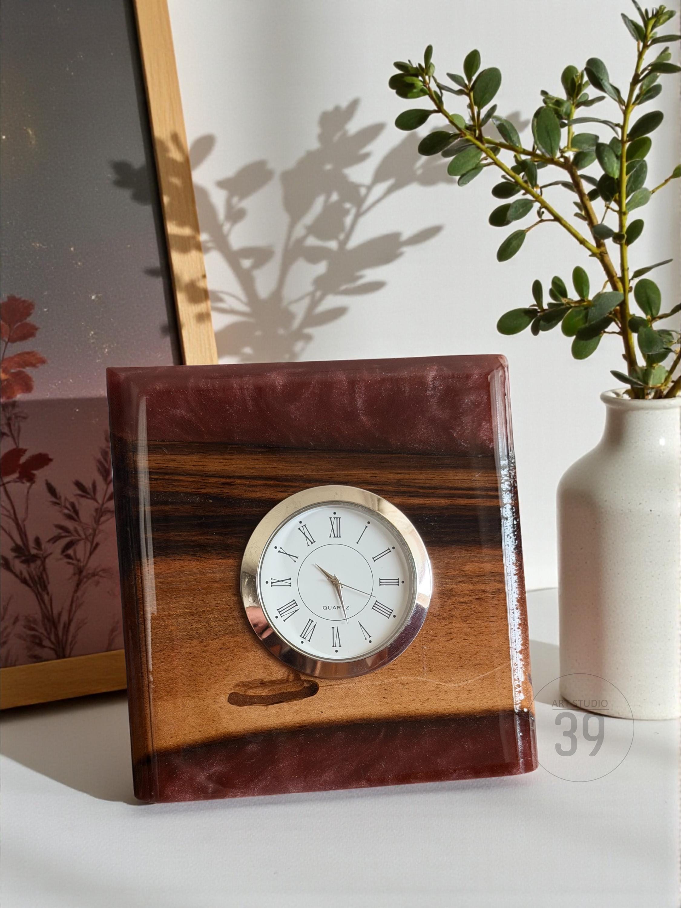 A square wooden clock with a silver-colored clock face and Roman numerals. The clock is set in a dark red resin frame. The wood grain is visible on the clock face. A small white vase with green leaves is to the right.
