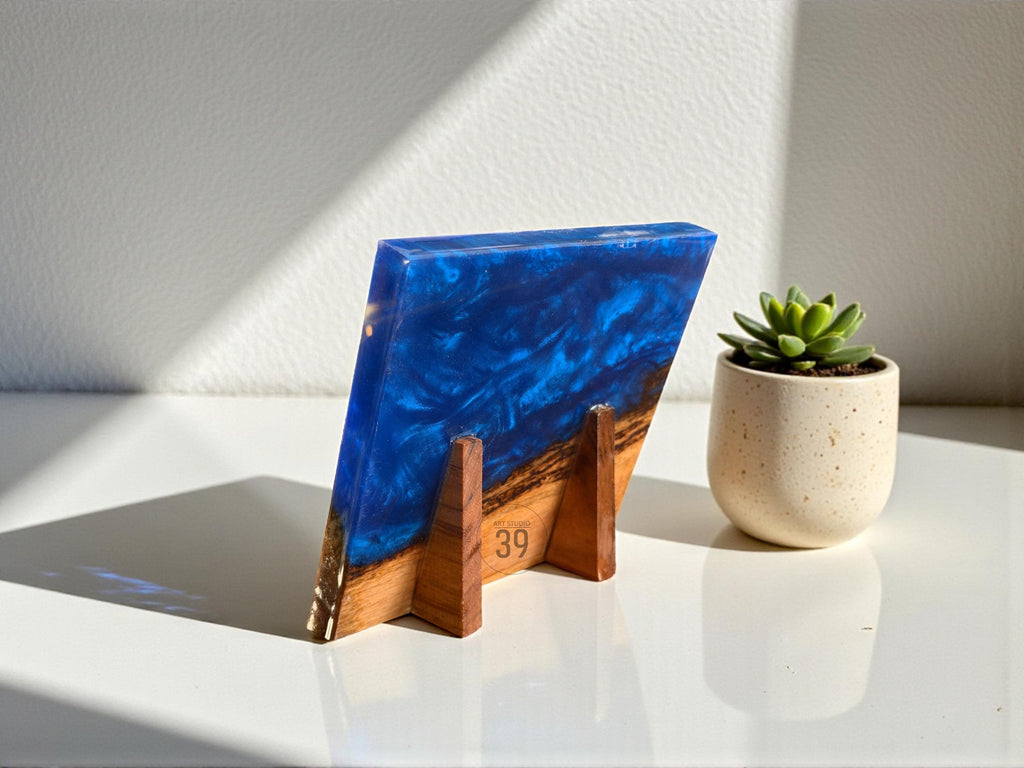 A rectangular resin art piece with a deep blue and white swirling design, supported by a wooden stand. The piece is angled, revealing a glimpse of a wooden base with the number "39" visible. A small succulent in a beige pot sits nearby.